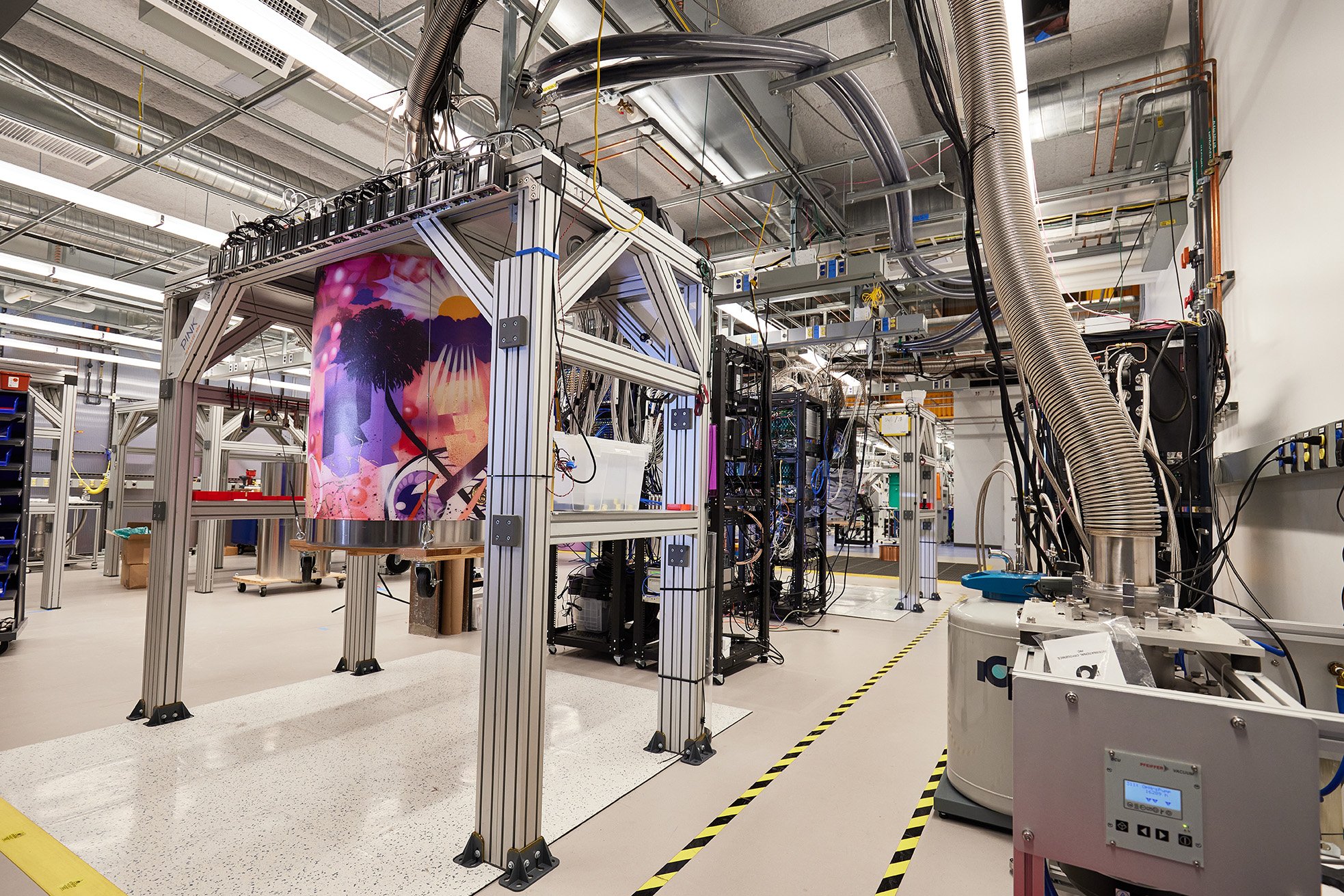 A large, complex piece of scientific equipment dominates a high-tech laboratory setting, with various wires, pipes, and support structures visible.