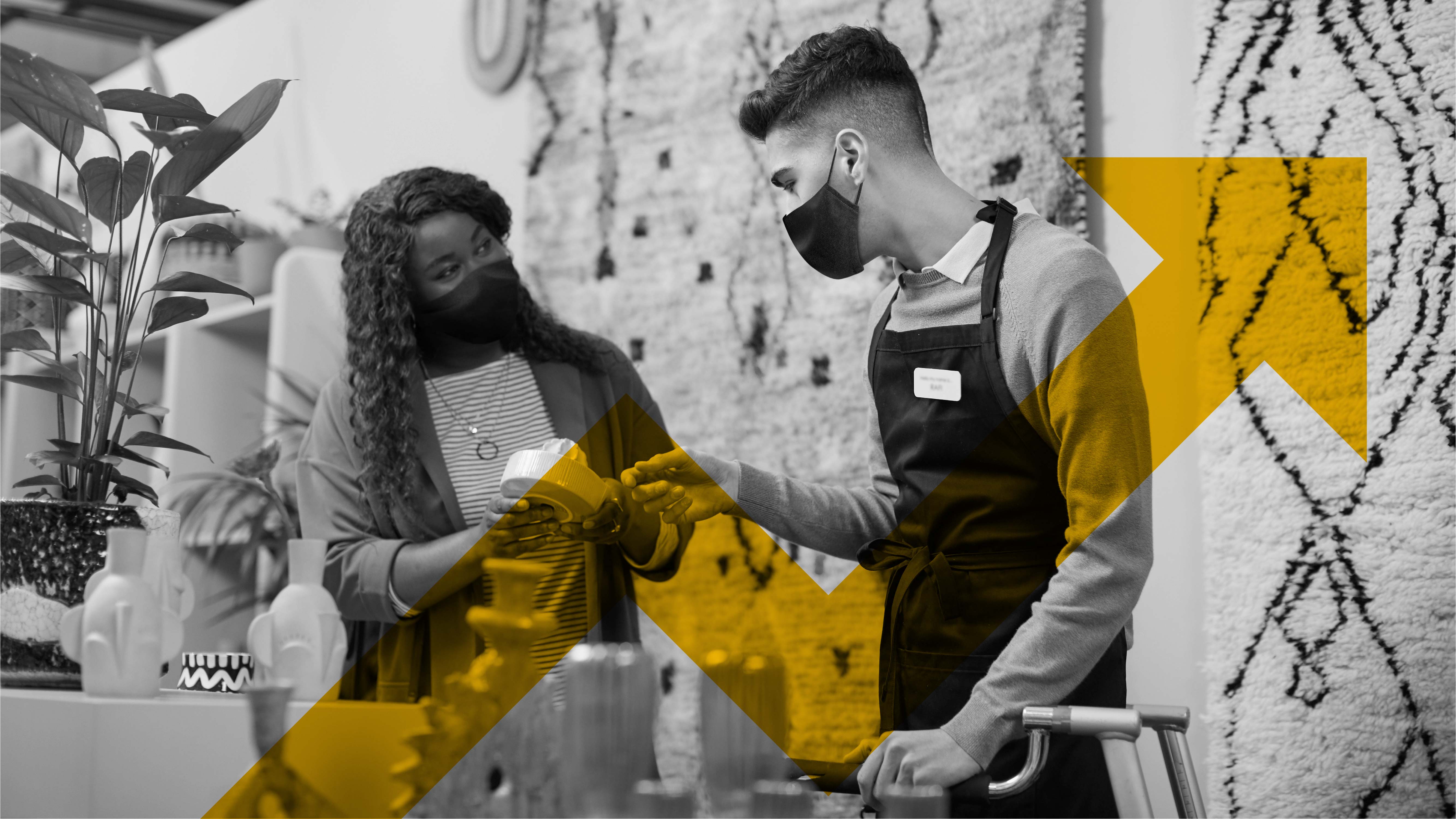A black-and-white photo of two individuals inside a store: a woman wearing a face mask speaks with a male employee in a face mask and shop apron. Overlaid on top of the image is a stylised translucent yellow tick pointing upwards.