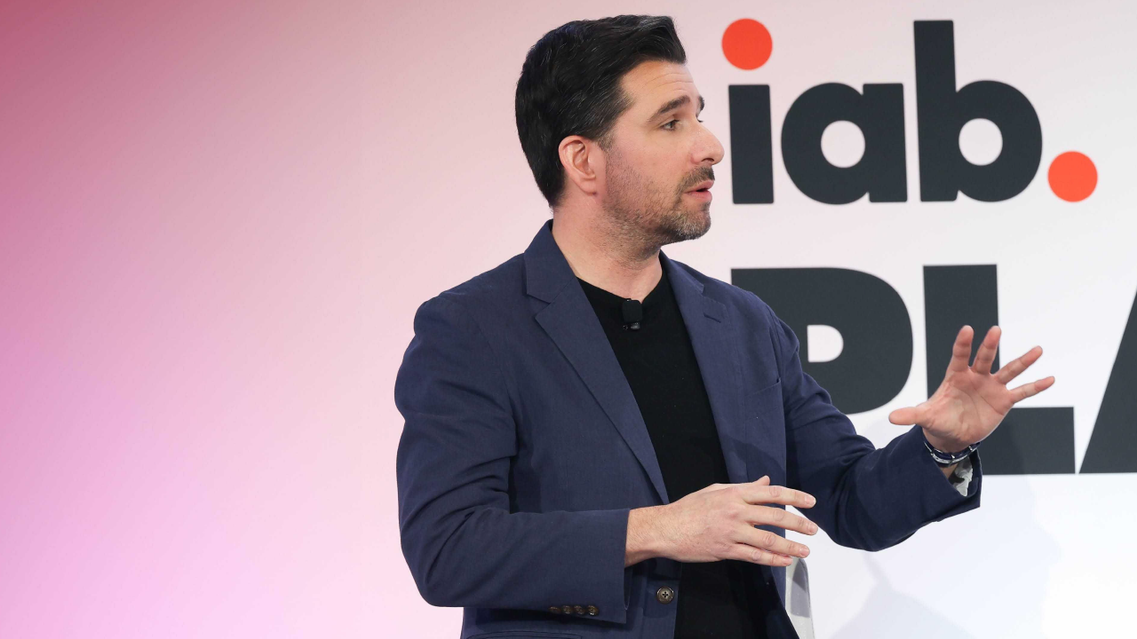 Jack Koch, SVP Research and Insights beim IAB, spricht auf einer Konferenzbühne. Koch sitzt auf einem hohen Hocker und gestikuliert mit seinen Händen. Er hat kurze dunkle Haare und trägt ein schwarzes Hemd, ein blaues Jackett und eine schwarze Hose.