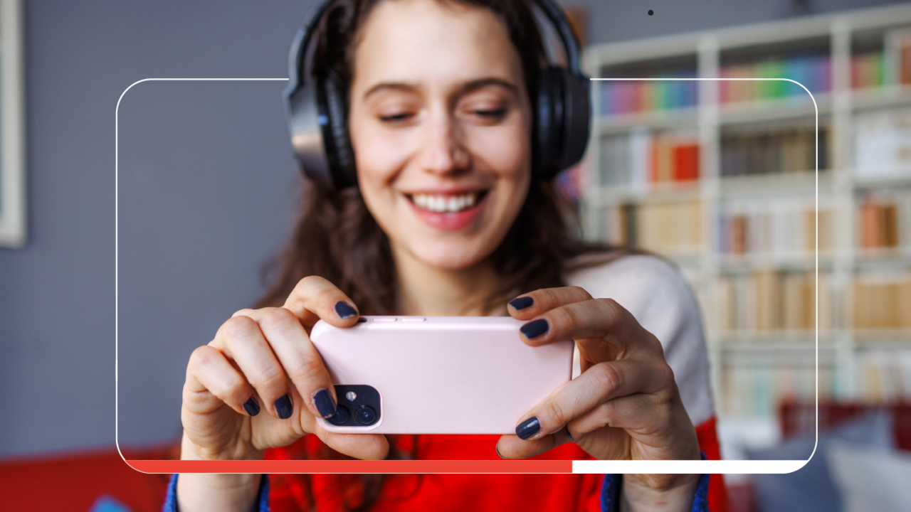 A woman with light skin and long dark hair wears a red and white top, black nail polish, and black over-the-ear headphones. She holds a mobile phone in a pink case on which she watches YouTube videos about her favorite games.