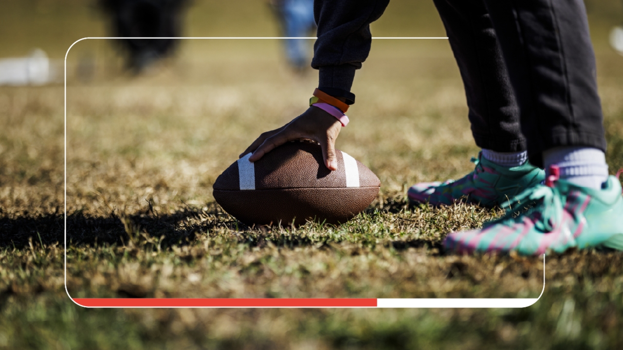 A football player in a black tracksuit and teal and pink cleats stands on the turf of a football field. Only a hand with dark skin and two legs and feet are in the frame. The hand holds the football at the feet, as if preparing to snap the ball.