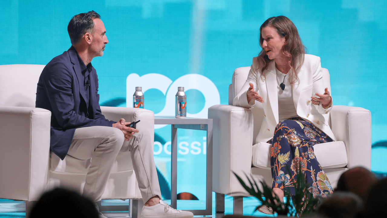 Sean Downey, president, Americas & Global Partners, Google, sits across from Laura Jones, CMO of Instacart, on the stage at Possible Miami in front of a teal and green background, decorated with a city skyline and palm trees.