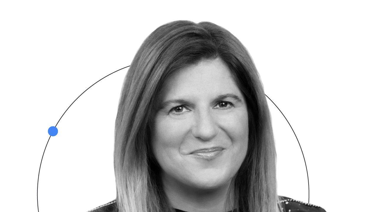 Google CMO Lorraine Twohill is pictured from the shoulders up, smiling. Twohill has light hair and light skin, and wears a black top. A line with a blue dot arcs behind her.