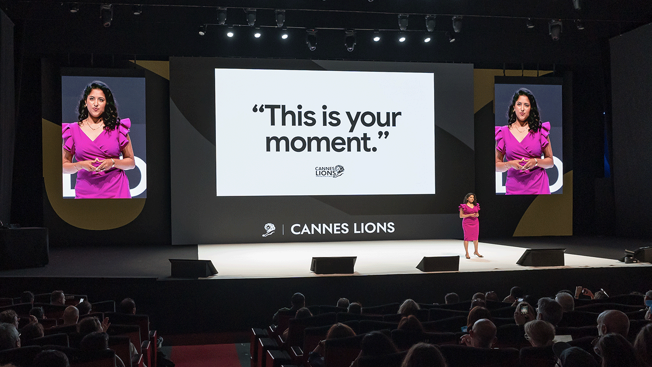 VP and GM of Google Ads Vidhya Srinivasan stands on the Palais Stage at Cannes delivering her keynote remarks. Wearing a hot pink dress, she stands in front of a huge screen that reads “This is your moment” between two monitors with her in close up.