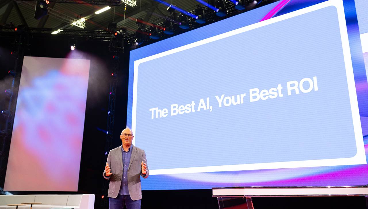Google'ın arama ve ticaret küresel reklam çözümlerinden sorumlu başkan yardımcısı Brendon Kraham, Köln’deki DMEXCO’da sahnede konuşuyor. Arkasında, soluk mavi bir ekranda beyaz harflerle “The Best AI, Your Best ROI” yazısı görünüyor.